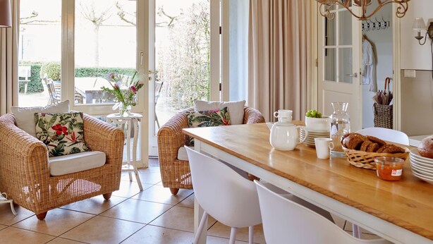 Een eetgedeelte met rieten meubels, een houten tafel en uitzicht op de tuin door grote glazen deuren.