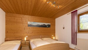 A wooden-paneled bedroom with two beds, a painting of mountains, a window with curtains, and a radiator.