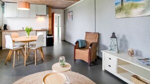 A modern kitchen with white cabinets, a dining table with chairs, a wicker chair, a small table with a glass vase, and a wall-mounted shelf with decorative items.