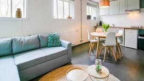 A modern apartment with a grey sofa, a dining table with chairs, a kitchen area, and a large window providing natural light.