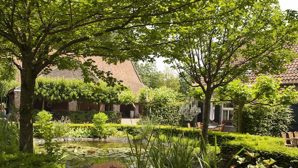 A serene garden with a pond, lush greenery, and a brick building with a tiled roof. - Holiday Home in Baarlo With Wood-fired Hot Tub (Baarlo)