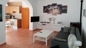 Una sala de estar con sofá, una mesa de centro y televisor. Se ve una cocina a través de un arco.