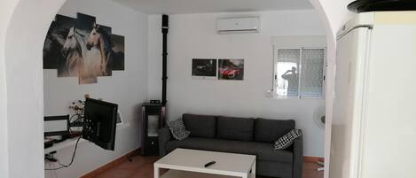 En stue med sofa, sofabord og fjernsyn. Væggene har indbyggede aircondition-anlæg og et vindue med persienner.
