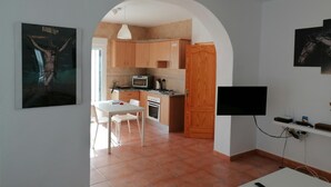 Una cocina moderna con armarios de madera, microondas y estufa. Hay una zona de comedor con una mesa y sillas blancas. Un televisor está montado en la pared.