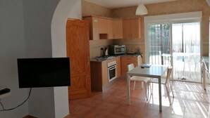 Una cocina compacta con armarios de madera, un microondas y una estufa a gas. Hay una mesa de comedor con sillas y un televisor montado en la pared.