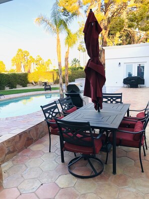 An outdoor patio with a pool, a table with chairs, and a grill.