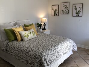 A neatly made bed with patterned bedding, a bedside lamp, and three framed botanical prints on the wall.
