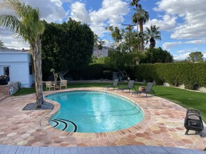 A swimming pool with a stone deck, surrounded by palm trees and a grassy area with lounge chairs.
