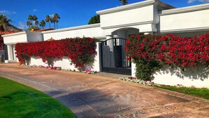 Una casa moderna con un camino de piedra que conduce a una entrada con puerta de rejas, rodeada de vegetación exuberante y flores rojas vibrantes.
