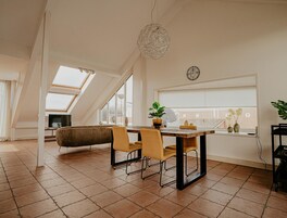 A modern loft apartment with a dining area featuring a wooden table and chairs, a large window with blinds, and a ceiling fan.