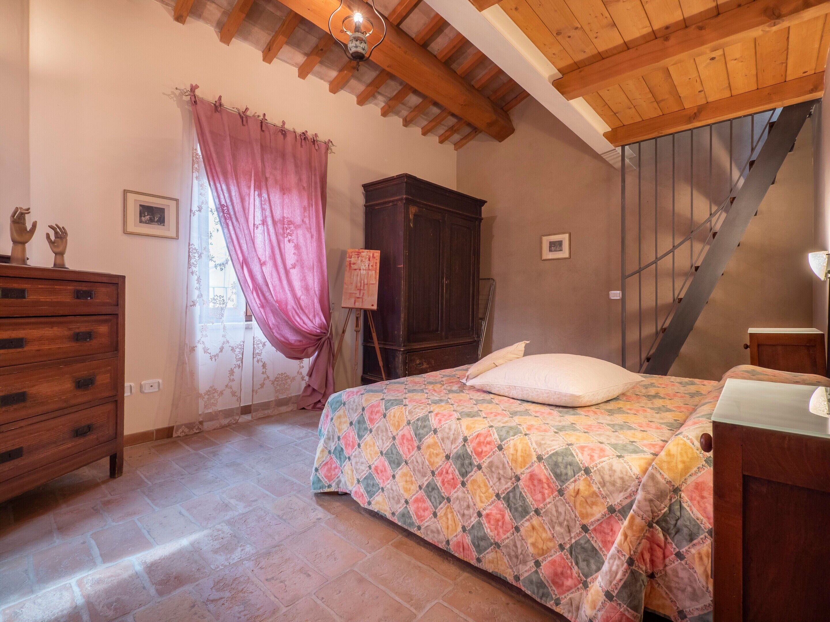 Une chambre avec une commode en bois, un lit recouvert d’une courtepointe aux couleurs vives, une armoire en bois et un escalier.