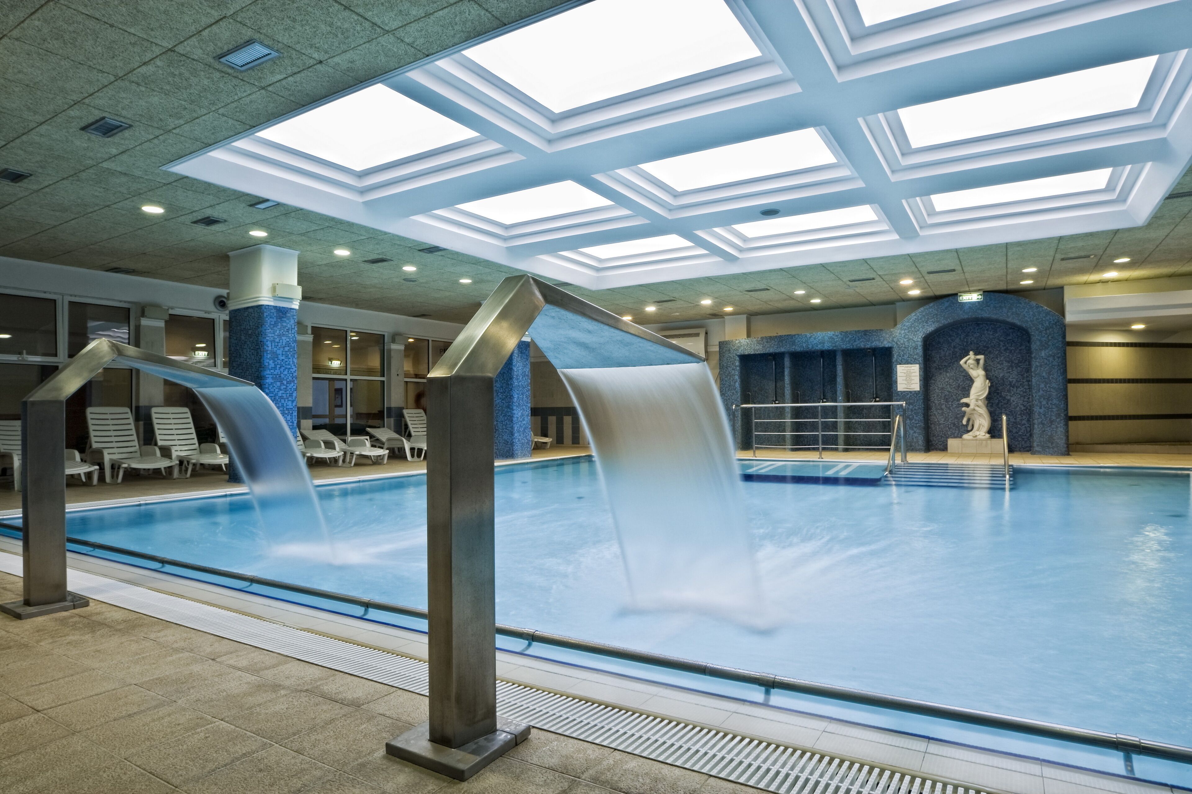 An indoor swimming pool with a waterfall feature, lounge chairs, and a statue.