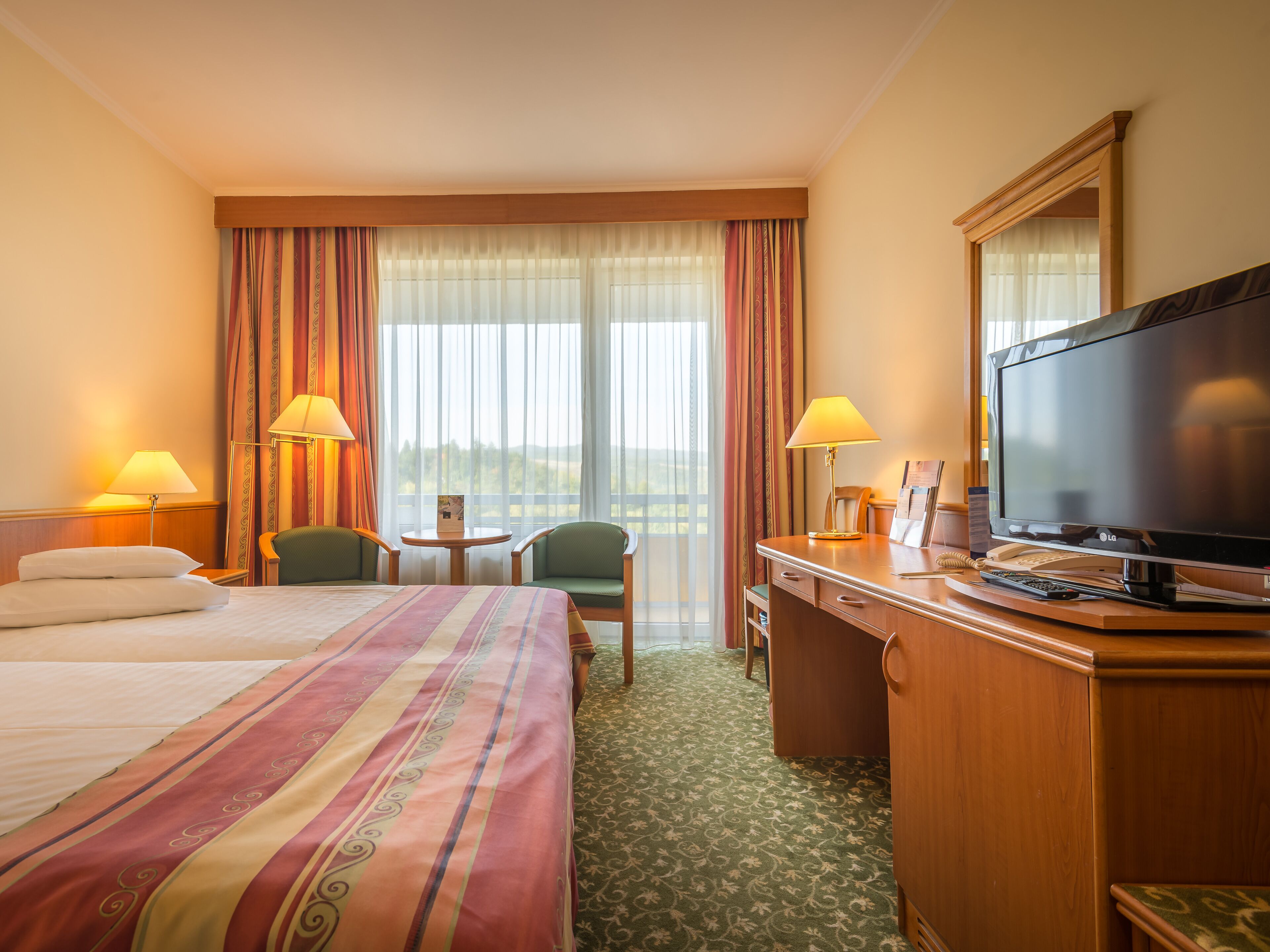 A hotel room with a bed, two chairs, a desk with a TV, and a view of the outdoors.