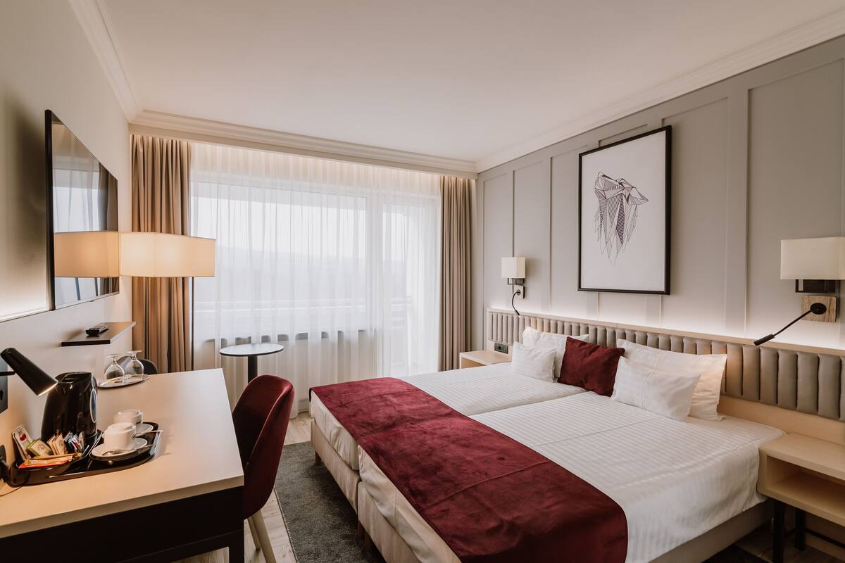 A hotel room with a bed, bedside tables, a desk with a lamp, a chair, and a window with curtains.