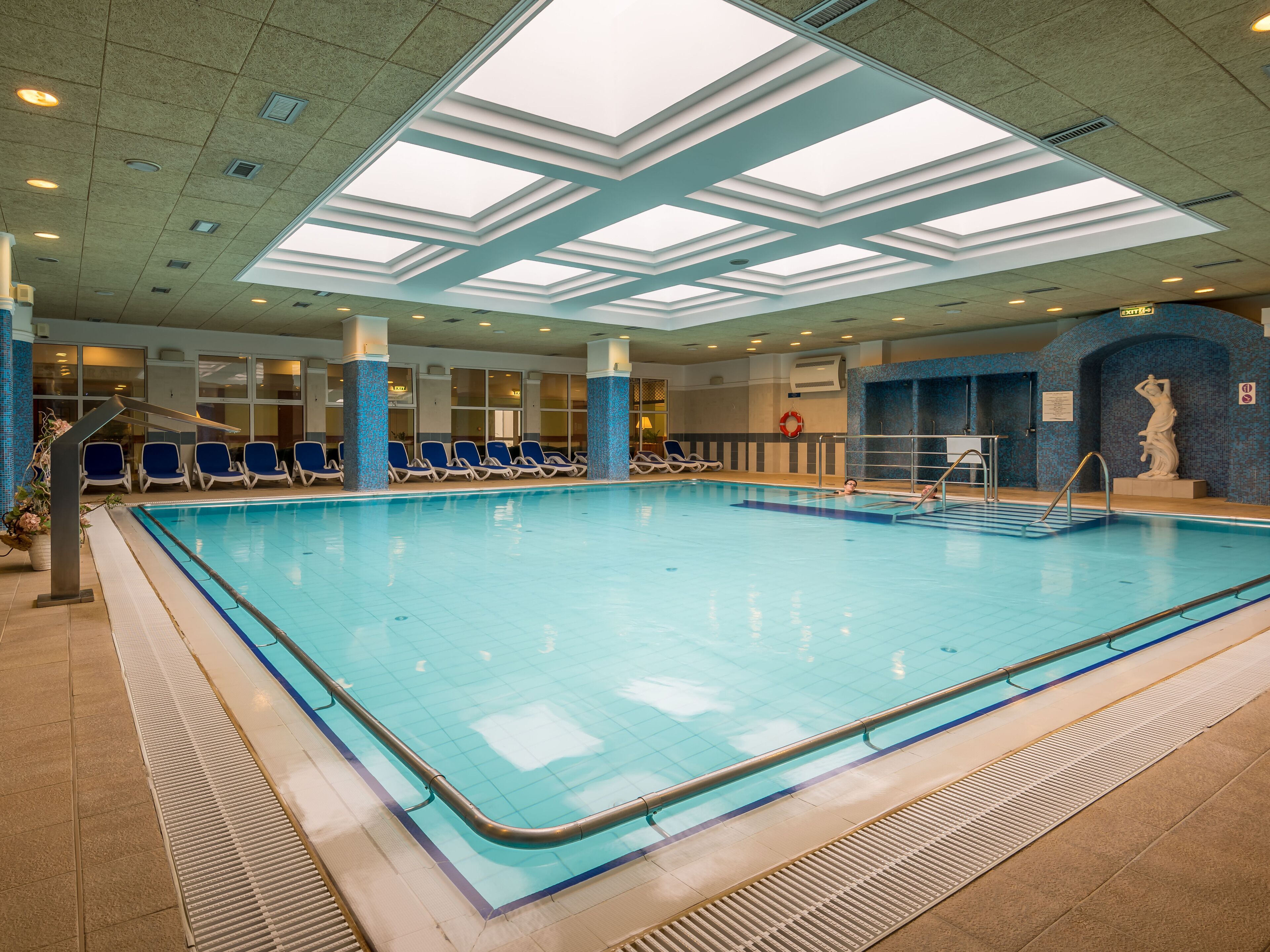 Ein Hallenbad mit klarem, blauem Wasser, umgeben von blauen Liegestühlen und einem gefliesten Boden.
