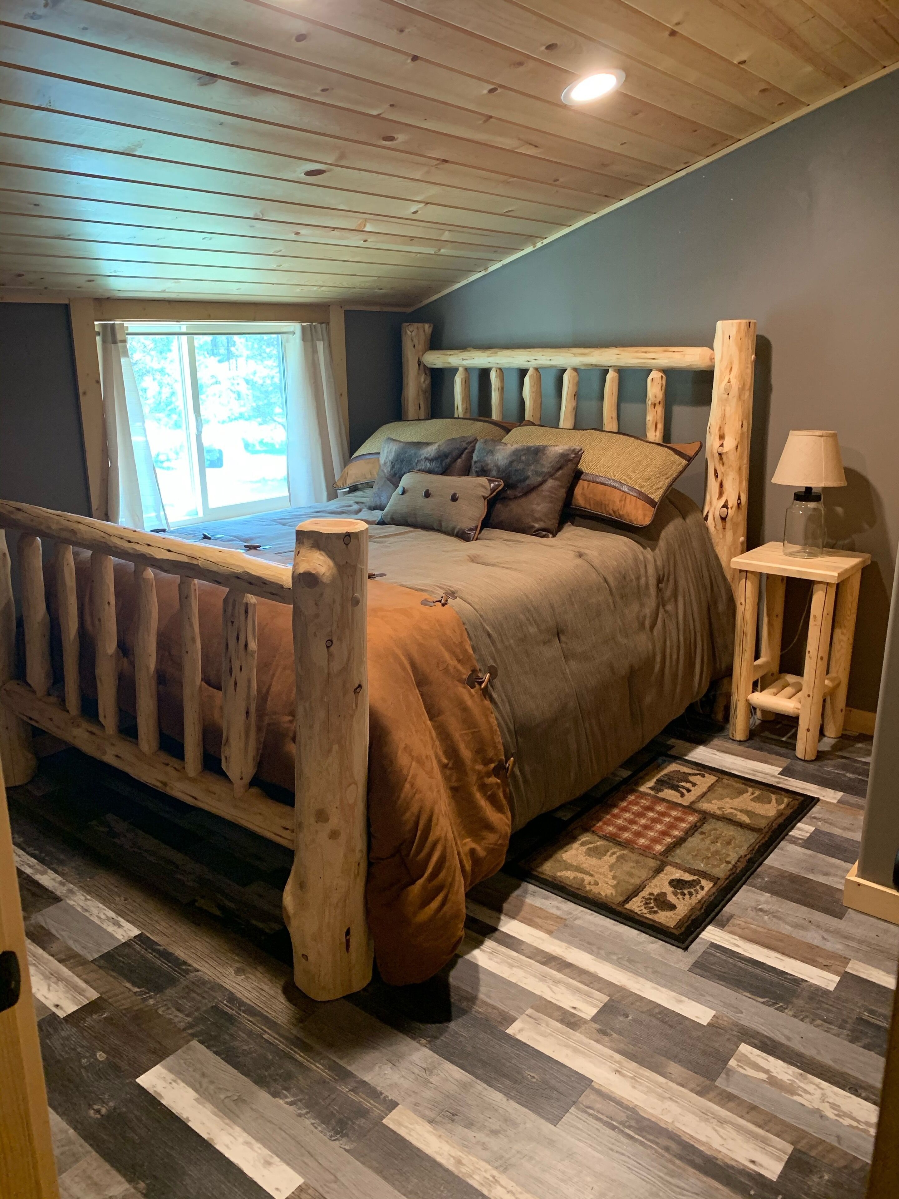 A bedroom with a wooden bed frame, a window with curtains, a bedside table with a lamp, and a patterned rug.