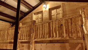Interior de una cabaña de madera con balcón, una lámpara de techo y una ventana con persiana de madera.
