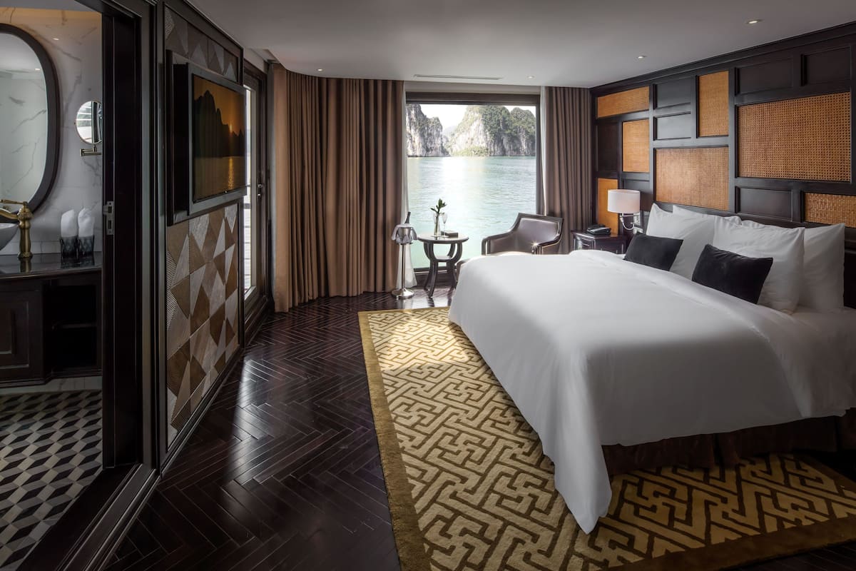 A hotel room with a large bed, a view of the water, a desk with a lamp, and a bathroom with a mirror and sink.