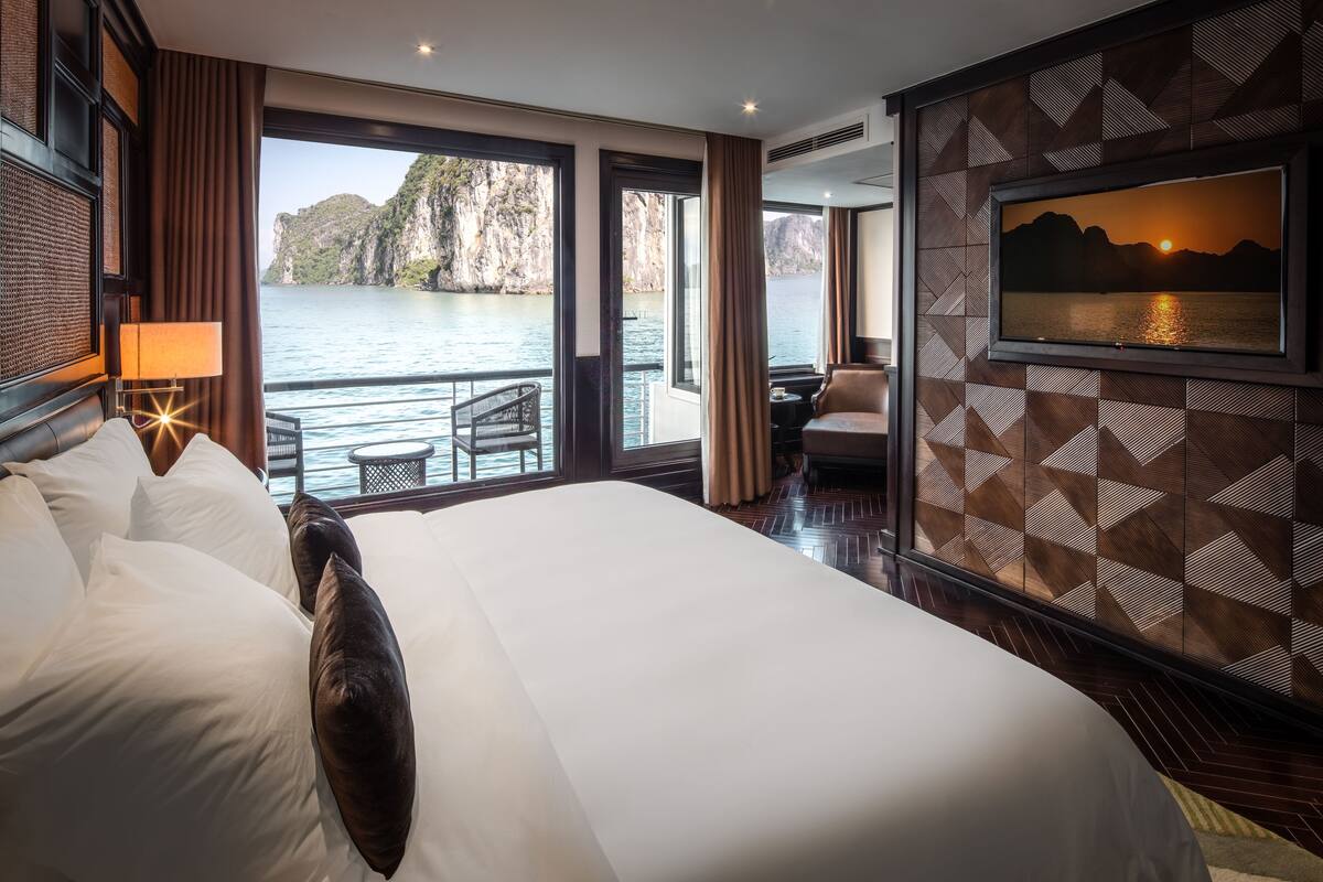 A hotel room with a large bed, a TV mounted on the wall, a balcony with a view of the water, and a dining area.