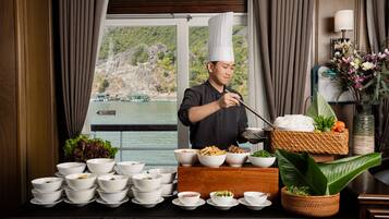 A chef preparing food at a buffet table with various dishes and a scenic view of a lake and mountains in the background.