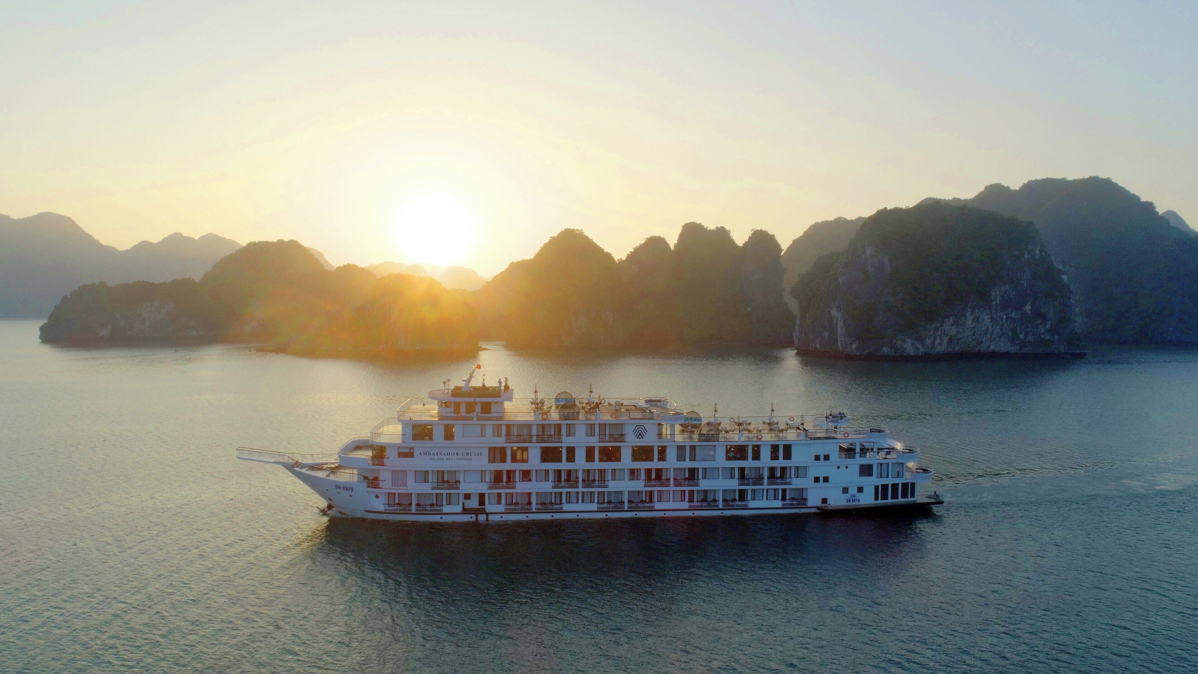 A large white cruise ship with multiple decks and a prominent bow, sailing in calm waters with a backdrop of rugged, forested islands under a setting sun.