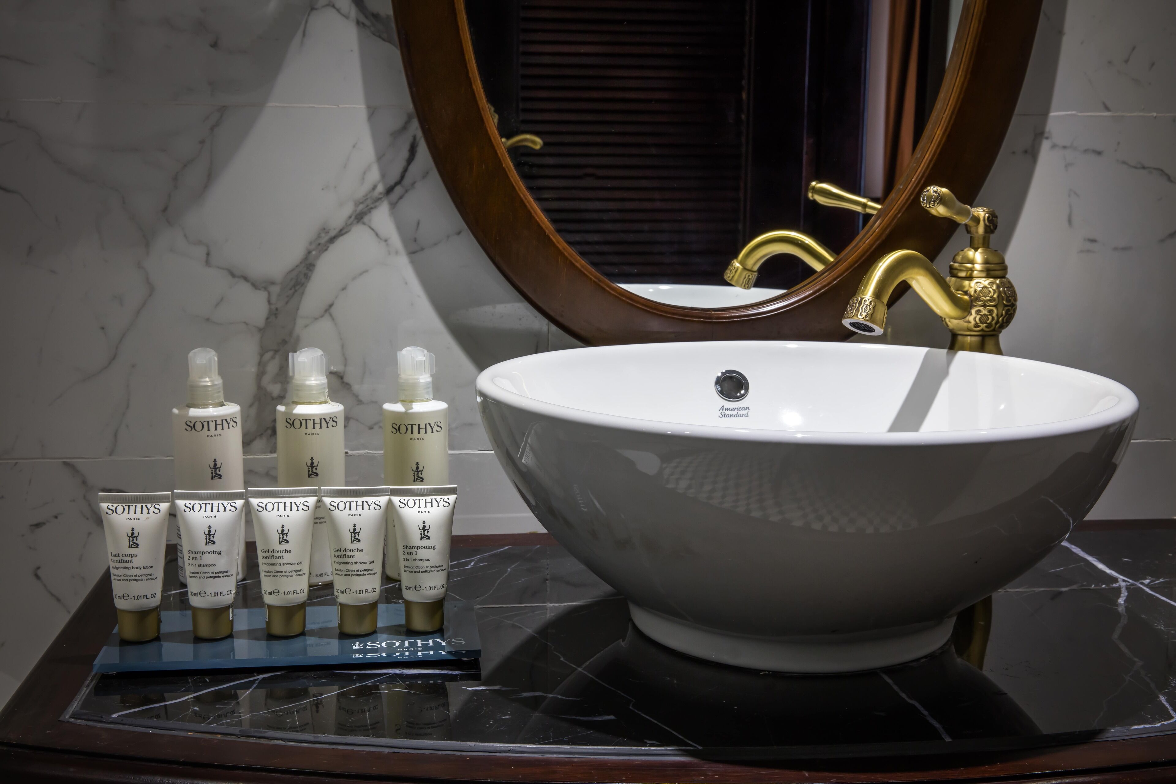 Un baño con un lavabo grande blanco, grifos dorados y una pared de mármol. Hay seis productos Sothys sobre una bandeja.