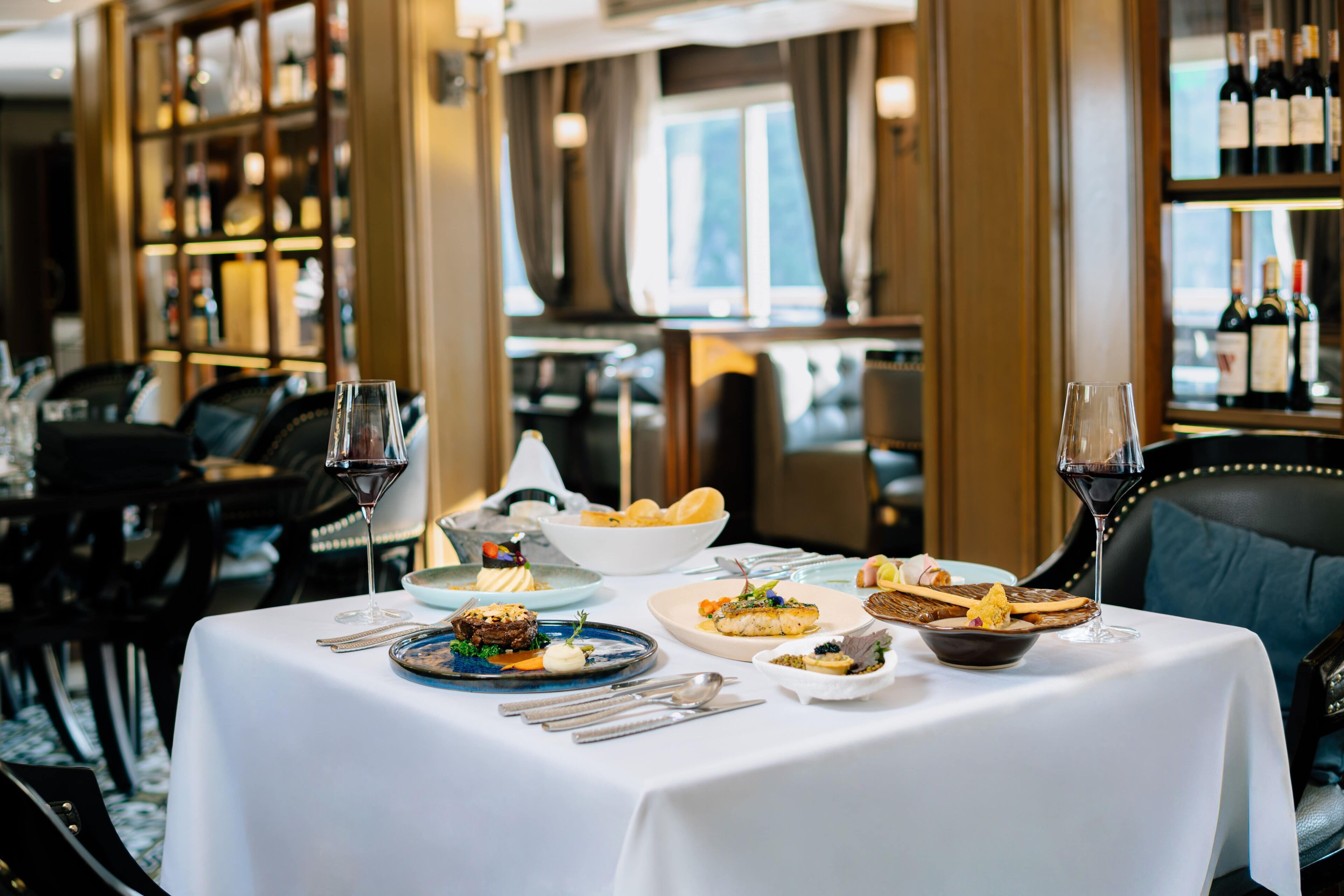 Una mesa de comedor preparada con diferentes platos, copas de vino y cubiertos, rodeada de sillas y una zona de bar con botellas.