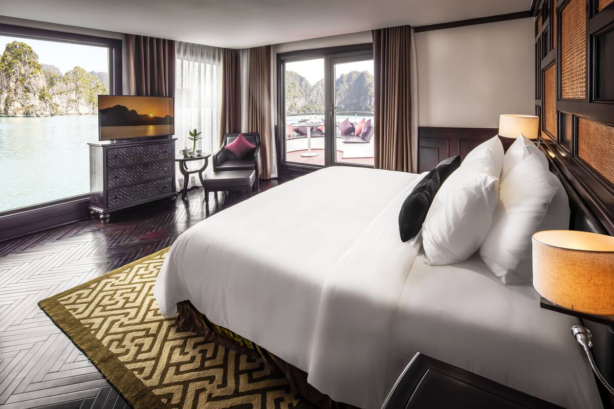 A large bed with white linens, a TV on a stand, a small table with a chair, and a view of a body of water through large windows.
