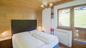 A bedroom with a wooden ceiling, a bed with white bedding, a nightstand with a lamp, and a window with a view of a grassy area. - Apartment With Panoramic View (Ramsau im Zillertal)