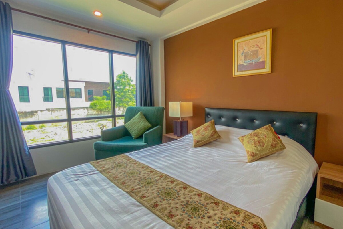 A hotel room with a bed, a chair, a nightstand, a lamp, a vase with flowers, and a window with curtains.