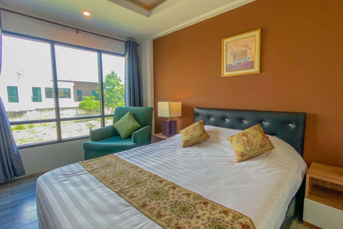 A hotel room with a bed, a chair, a nightstand, a lamp, a vase with flowers, and a window with curtains.