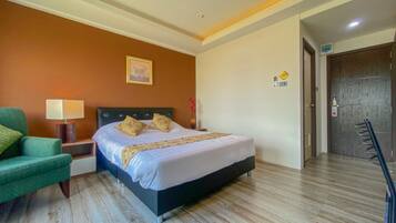 A hotel room with a bed, a green armchair, a nightstand, a lamp, and a door.