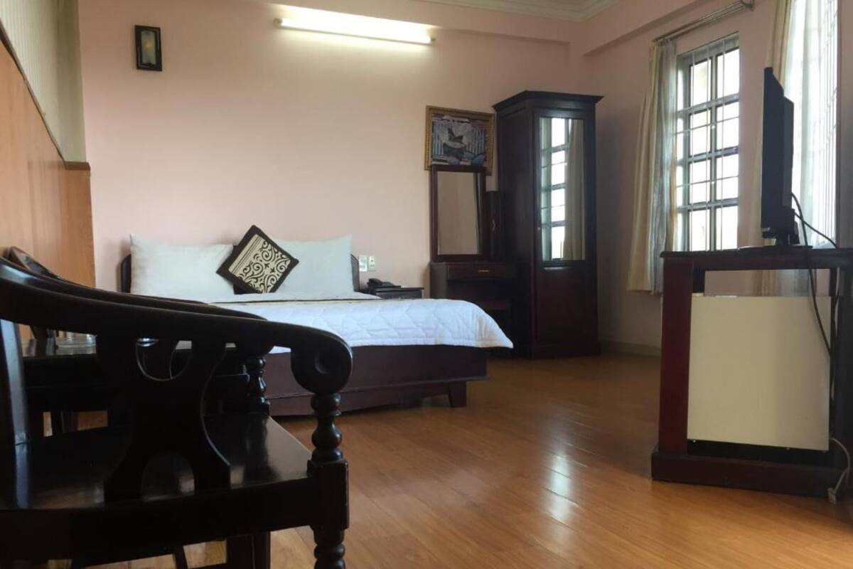 A bedroom with a bed, a wooden chair, a desk with a computer, a window, and a door.