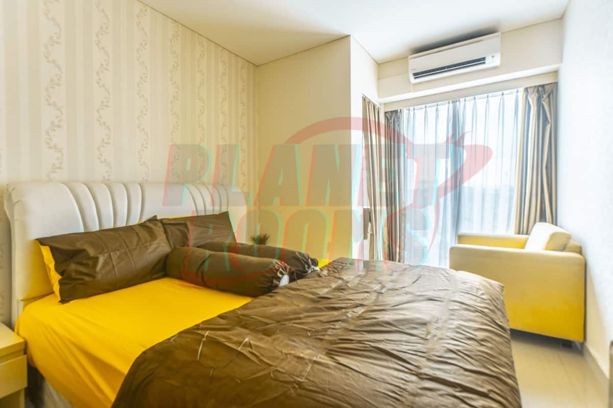 Une chambre d’hôtel avec un grand lit, une couvre-lit jaune, une couverture marron, une petite plante en pot, un fauteuil jaune et une fenêtre avec des rideaux.