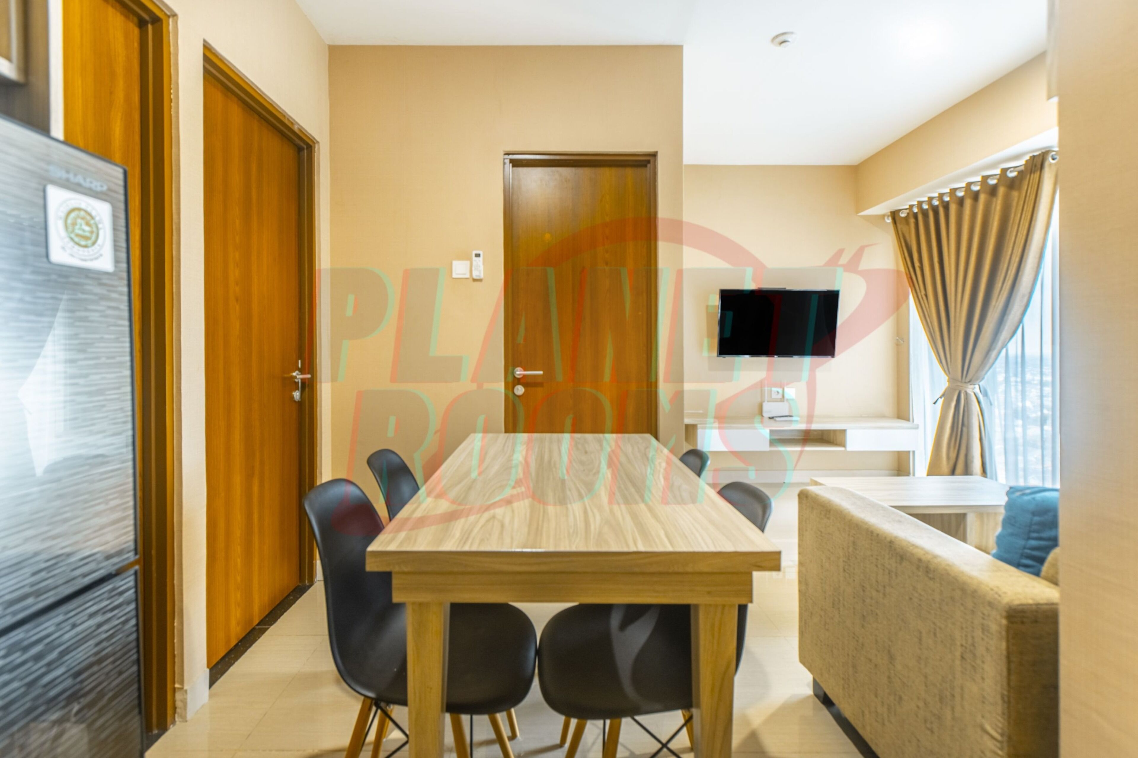 A modern hotel room with a wooden dining table, black chairs, a sofa, a TV, and a window with curtains.