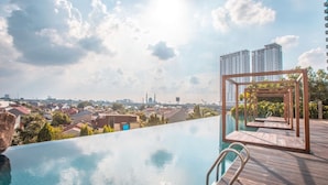 A rooftop infinity pool with a wooden deck and a view of a cityscape, including high-rise buildings and a mosque. - 8rooms@ Grand Kamala Lagoon (Bekasi)