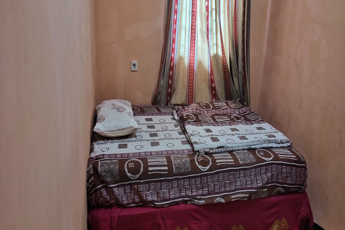 A small, narrow room with a bed covered in patterned bedding, a decorative pillow, and a curtain with a red and beige pattern.