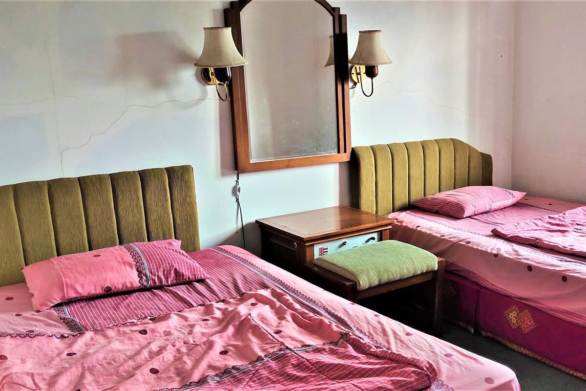 Une chambre avec deux lits, chacun agrémenté de draps et d’oreillers roses, une table de chevet en bois et un miroir avec des lampes fixées au mur.