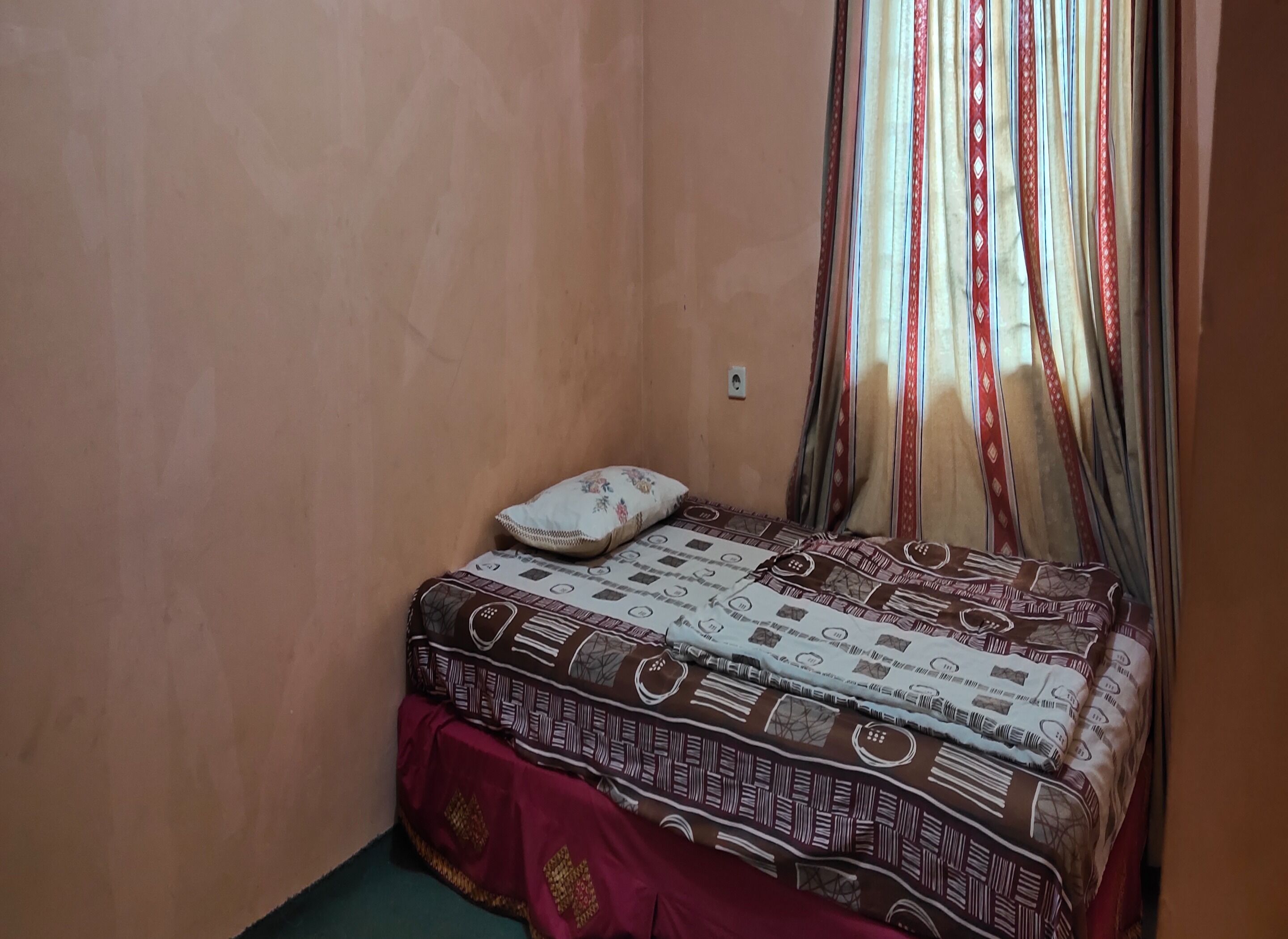 A small, single-bed room with a patterned bedspread, a pillow, and curtains.