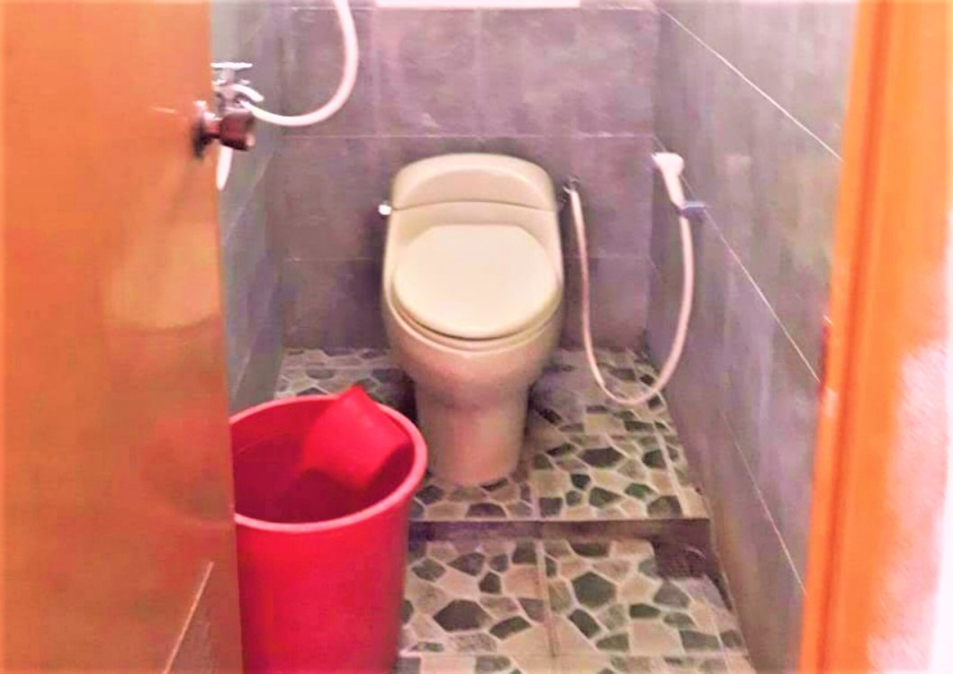 Un baño pequeño con inodoro, un cubo rojo y suelo de baldosas.