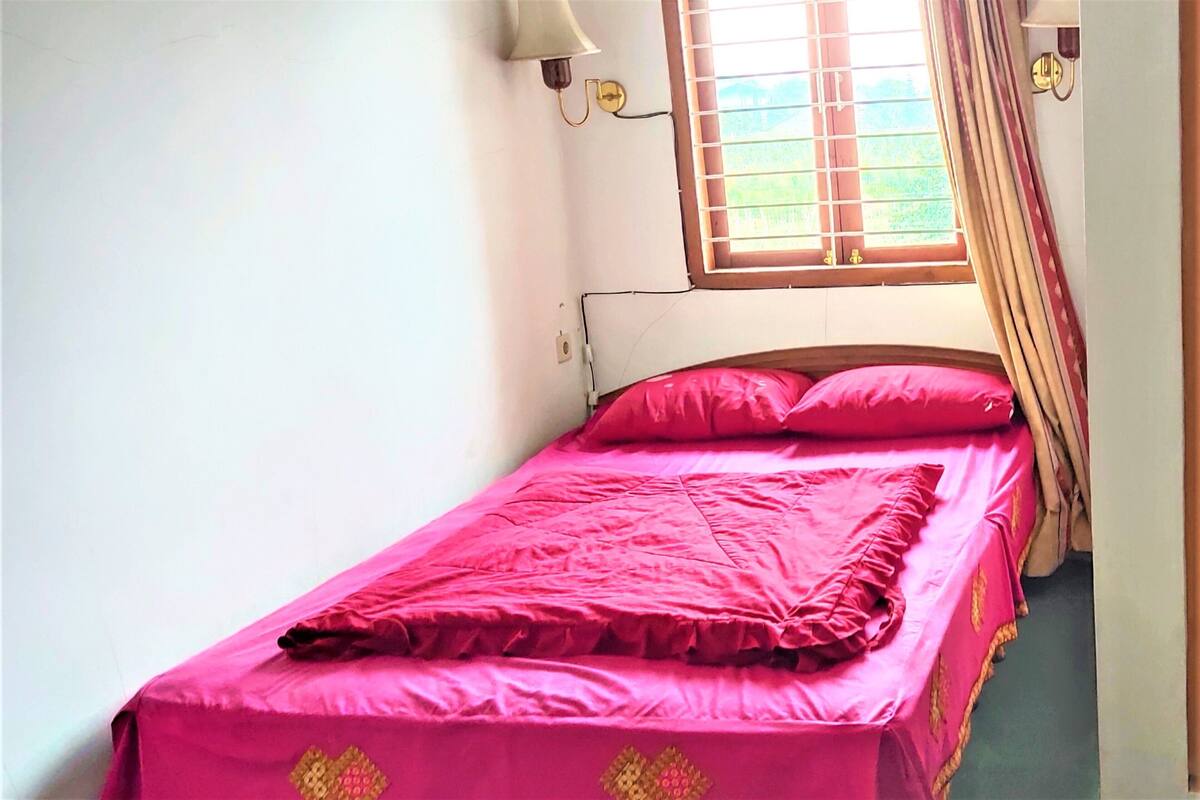 Un lit simple avec une couvre-lit et des oreillers roses, une fenêtre avec des rideaux et un luminaire fixé au mur.