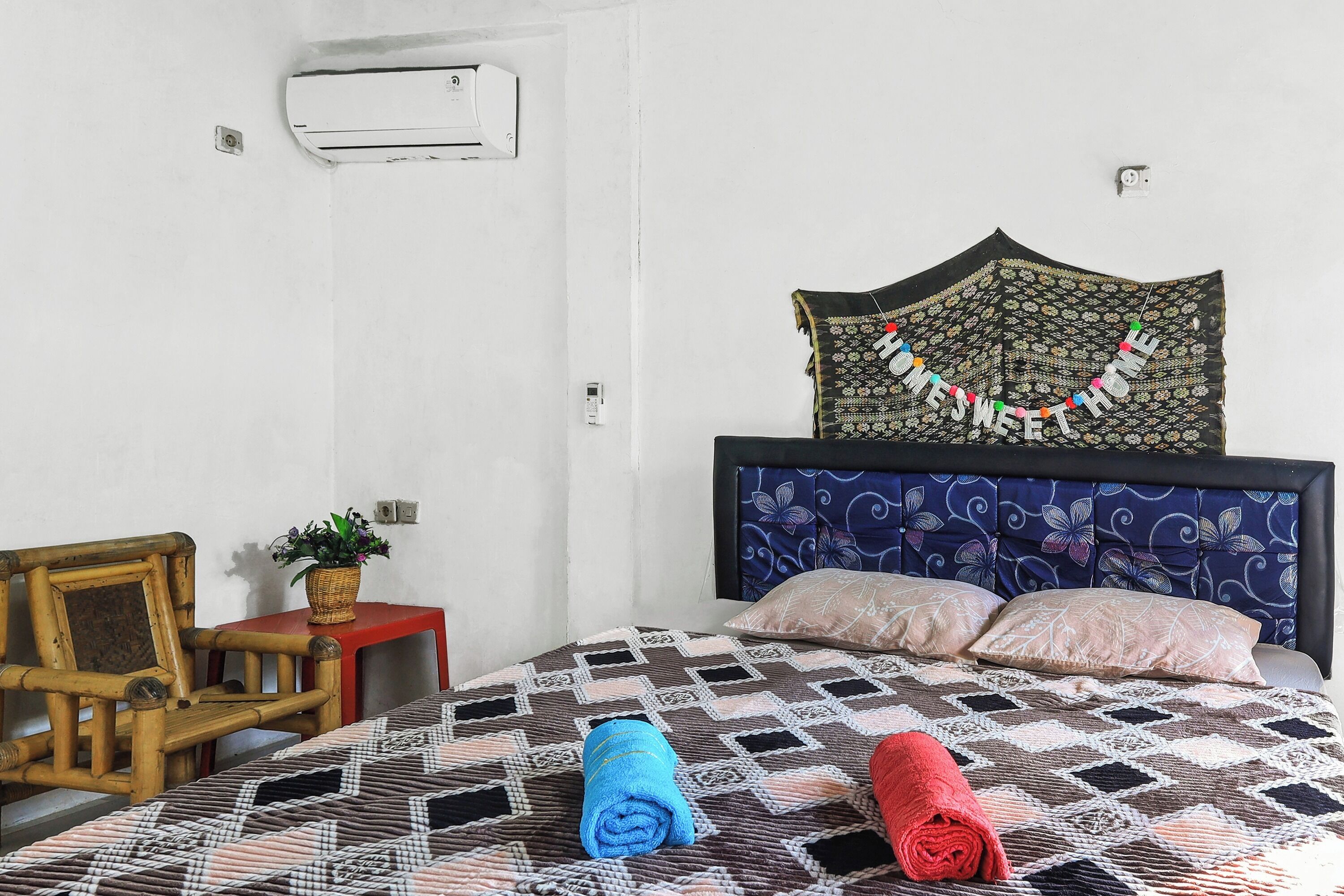 A bedroom with a bed, a headboard, a wall-mounted air conditioner, a small table with a plant, and a chair.