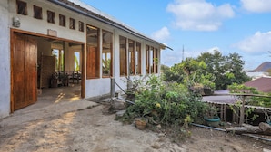 Un bâtiment blanc avec des portes et des fenêtres en bois, un porche couvert et un chemin de terre menant à un banc en bois.