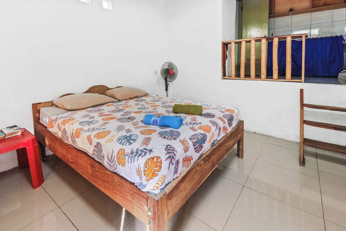 Een slaapkamer met een houten bed, een rode kruk, een ventilator en een raam met een blauw gordijn.