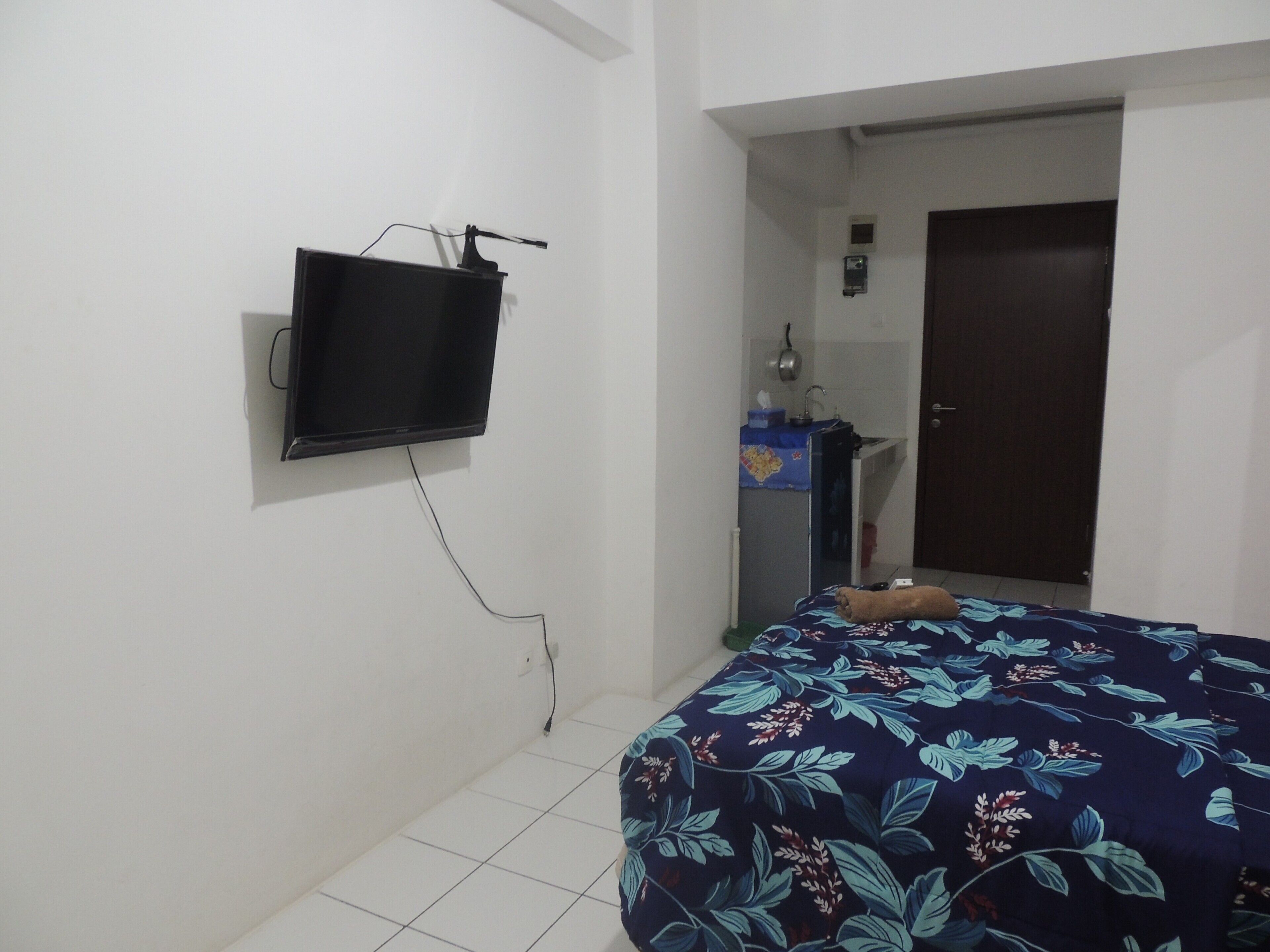 A bedroom with a bed, a TV mounted on the wall, a door, and a small kitchenette area.