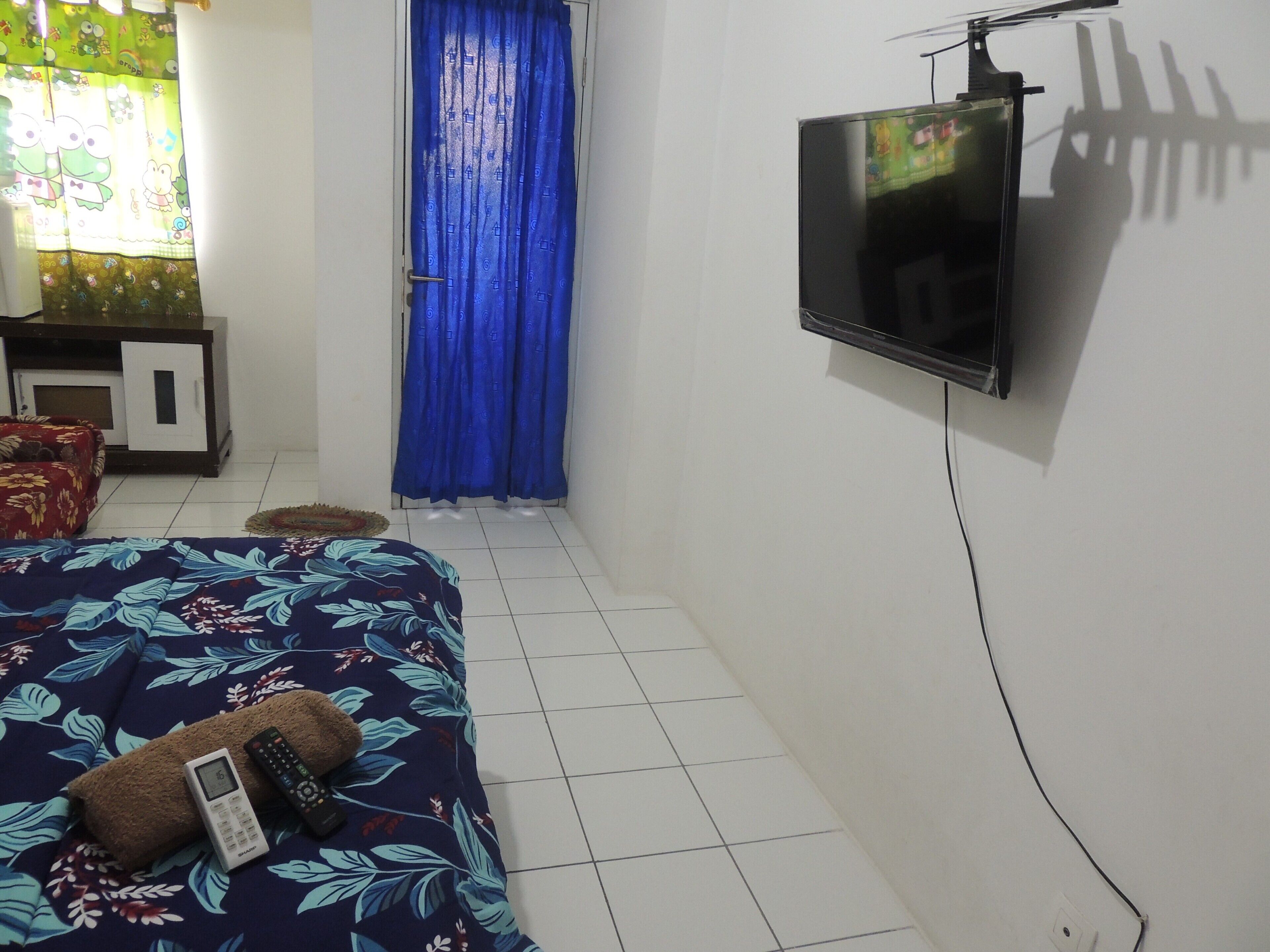 A bedroom with a bed, a TV mounted on the wall, a small cabinet, and a window with curtains.