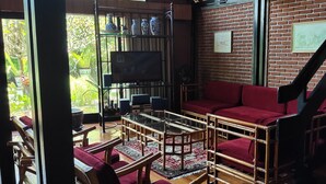 A cozy living area with a red sofa, a glass coffee table, and a TV on a shelf. There are potted plants and a brick wall in the background. - OBC Villa (Bandung)