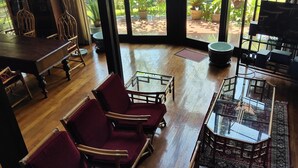 A spacious room with a wooden floor, a dining table with chairs, a seating area with red chairs, a glass coffee table, and a view of a garden through large windows. - OBC Villa (Bandung)