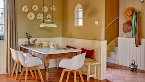 A dining area with a wooden table, white chairs, and a bench. There are hanging plates on the wall, a window, and a staircase with a hat and bag hanging on the wall.