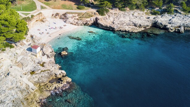 Ett flygbildsfoto av ett kustområde med en strand, en klippig kustlinje och klart turkosfärgat vatten.